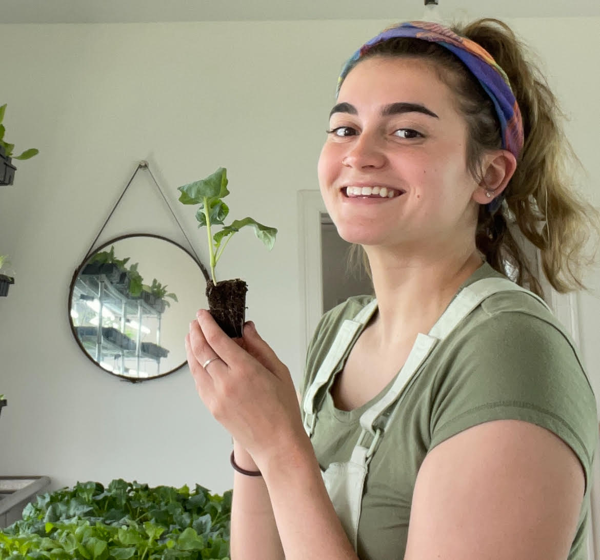 A person smiling while holding a small plant in a room with more plants and a mirror.