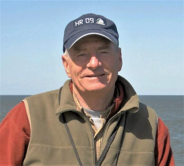 An older man in a baseball cap and jacket stands outdoors near water under a clear sky.