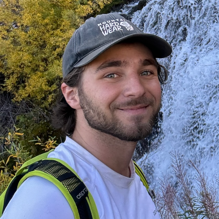 A person with a backpack and cap smiles near a waterfall with greenery around.