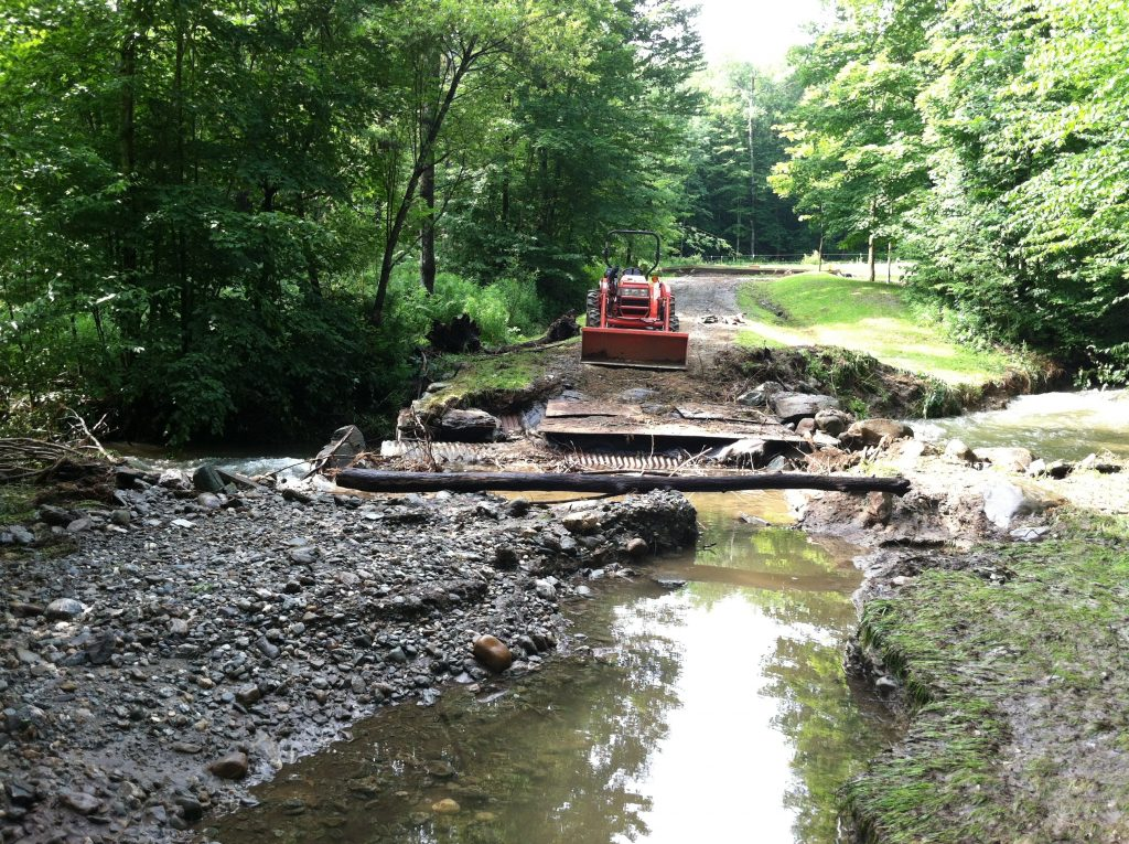A damaged dirt road with debris and a small stream, a tractor in the background, surrounded by dense green forest.
