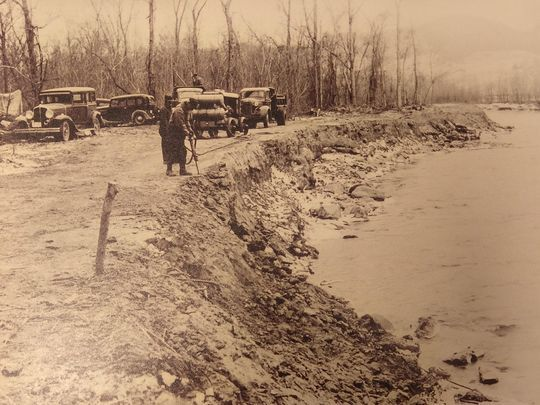 A group of vintage cars parked near a riverbank, with people standing and trees in the background.