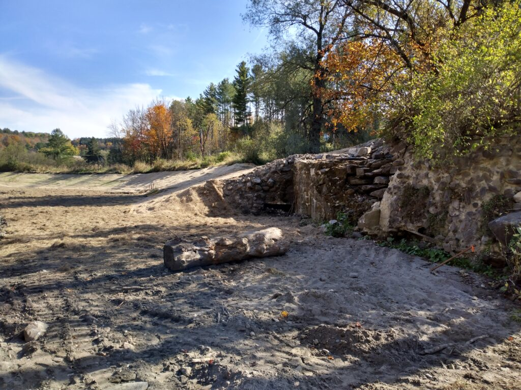 A sandy clearing with a log, trees with autumn foliage, and a stone wall in the background under a blue sky.
