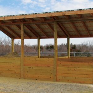 A wooden structure with a slanted roof, possibly a storage or shelter area, with a pile of material inside and a backdrop of trees.