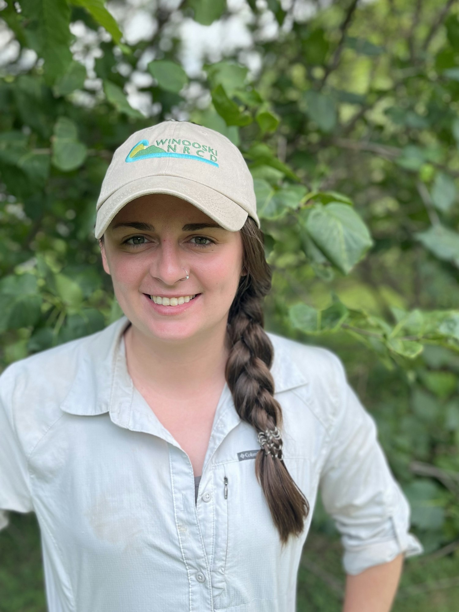 Person outdoors wearing a "Winooski NRCD" cap, long braid, and a light-colored shirt, smiling.