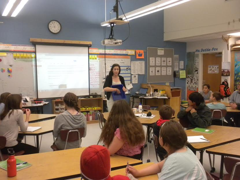 A teacher is standing in front of a classroom with students seated at tables, a projector screen, and educational materials on the walls.