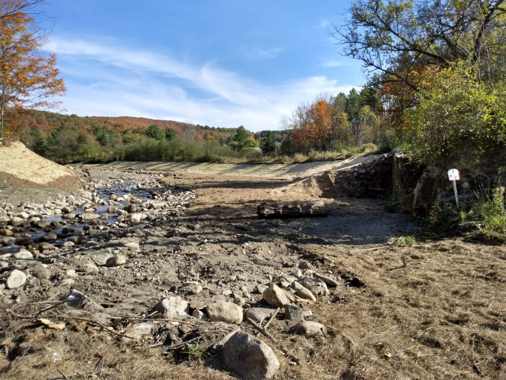 Rocky streambed with a small creek, surrounded by autumn trees and a clear blue sky.