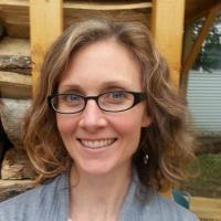 A person with glasses smiling, standing in front of a stack of wood logs.