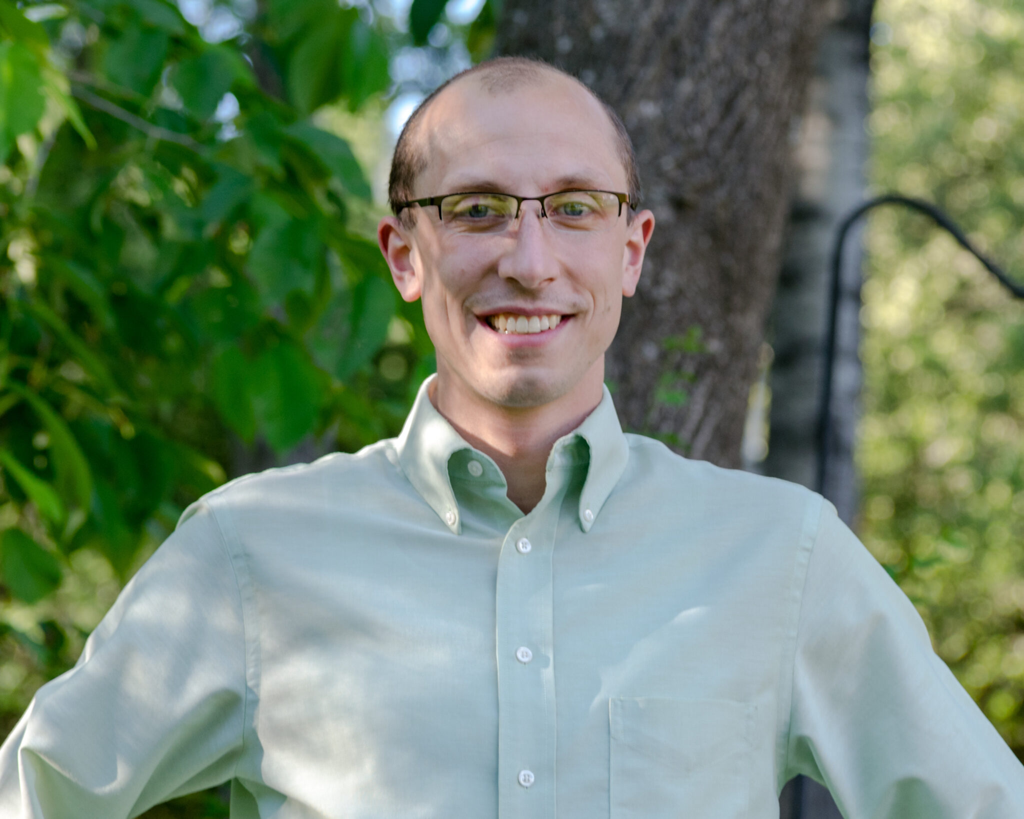 A person smiling, wearing glasses and a light green shirt, standing outdoors with green leaves and a tree in the background.