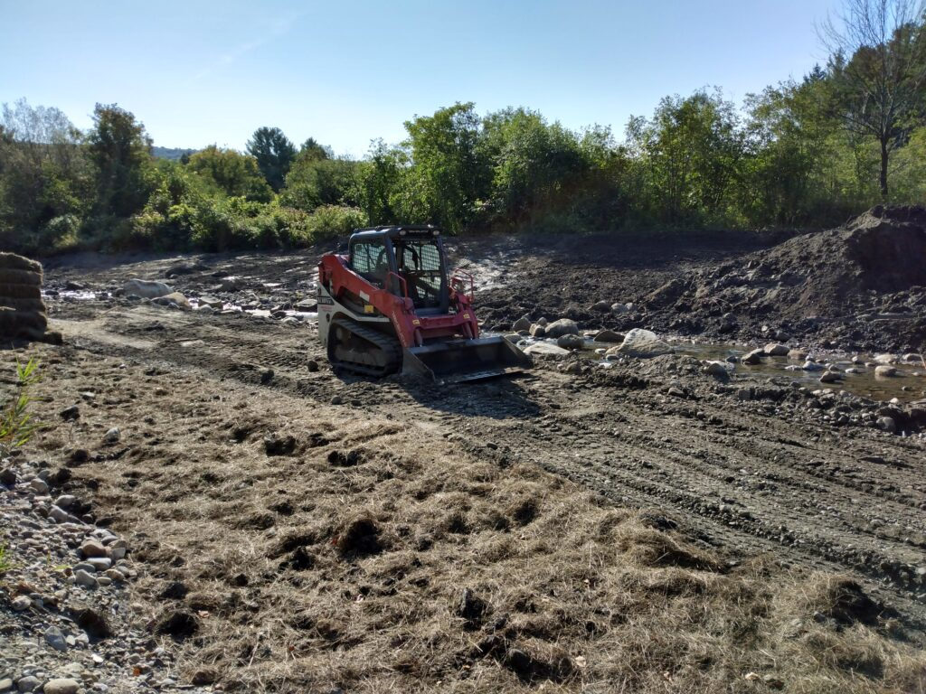 A red bulldozer on a dirt path near a small stream with trees in the background.
