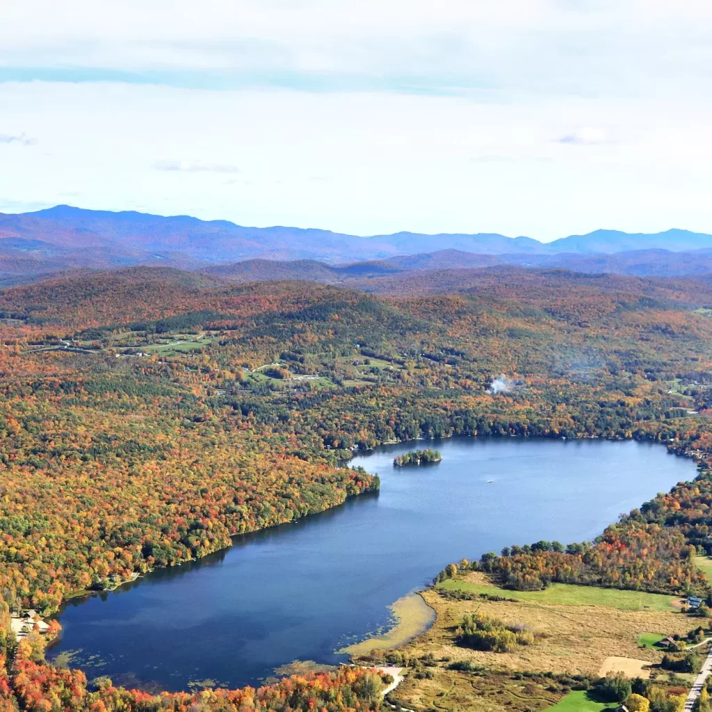 Aerial view of a scenic lake surrounded by colorful autumn forests and distant rolling hills under a clear sky.