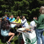 A group of people is seated outdoors at a table, listening to a person speaking next to a table with materials and a "Waste" sign.