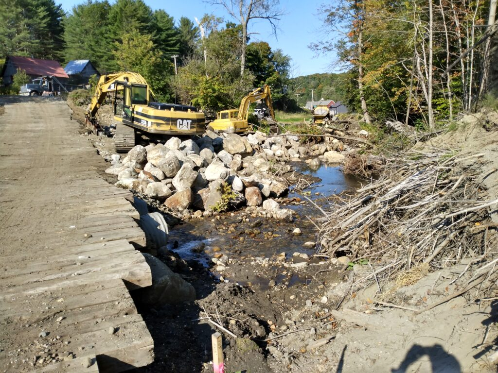 Construction site with excavators near a creek, rocks, and a dirt path.