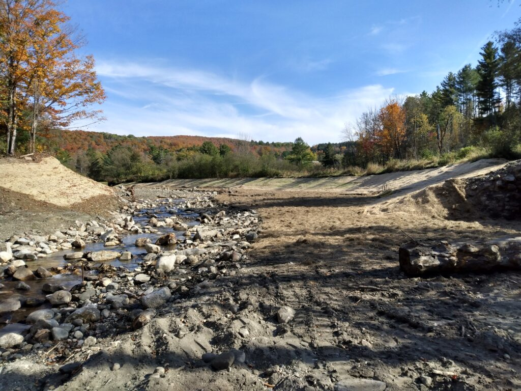 Rocky riverbed with sparse water flow, surrounded by trees with autumn foliage under a blue sky.