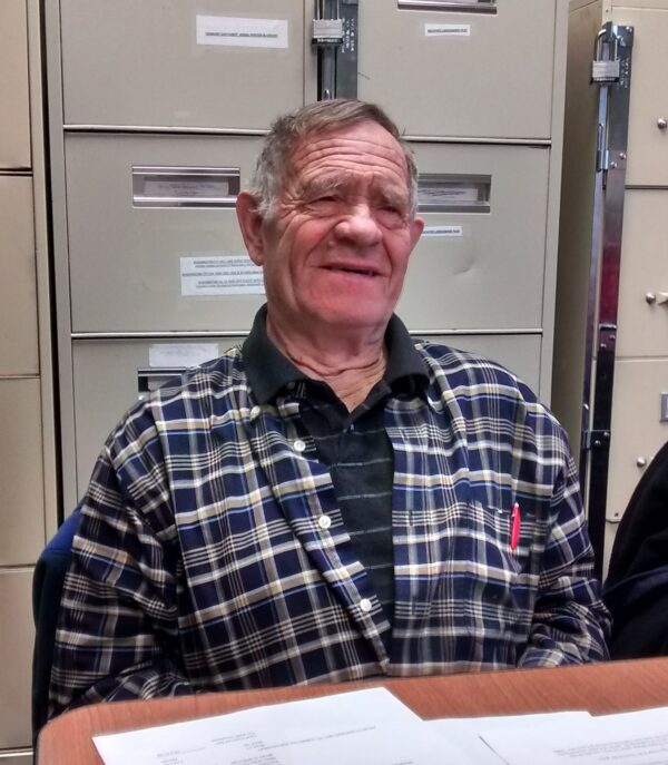An older man in a checkered shirt sits at a table with papers, in front of filing cabinets.