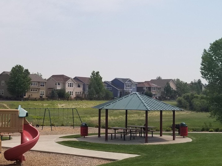A playground with a slide, swings, a gazebo with picnic tables, and houses in the background.
