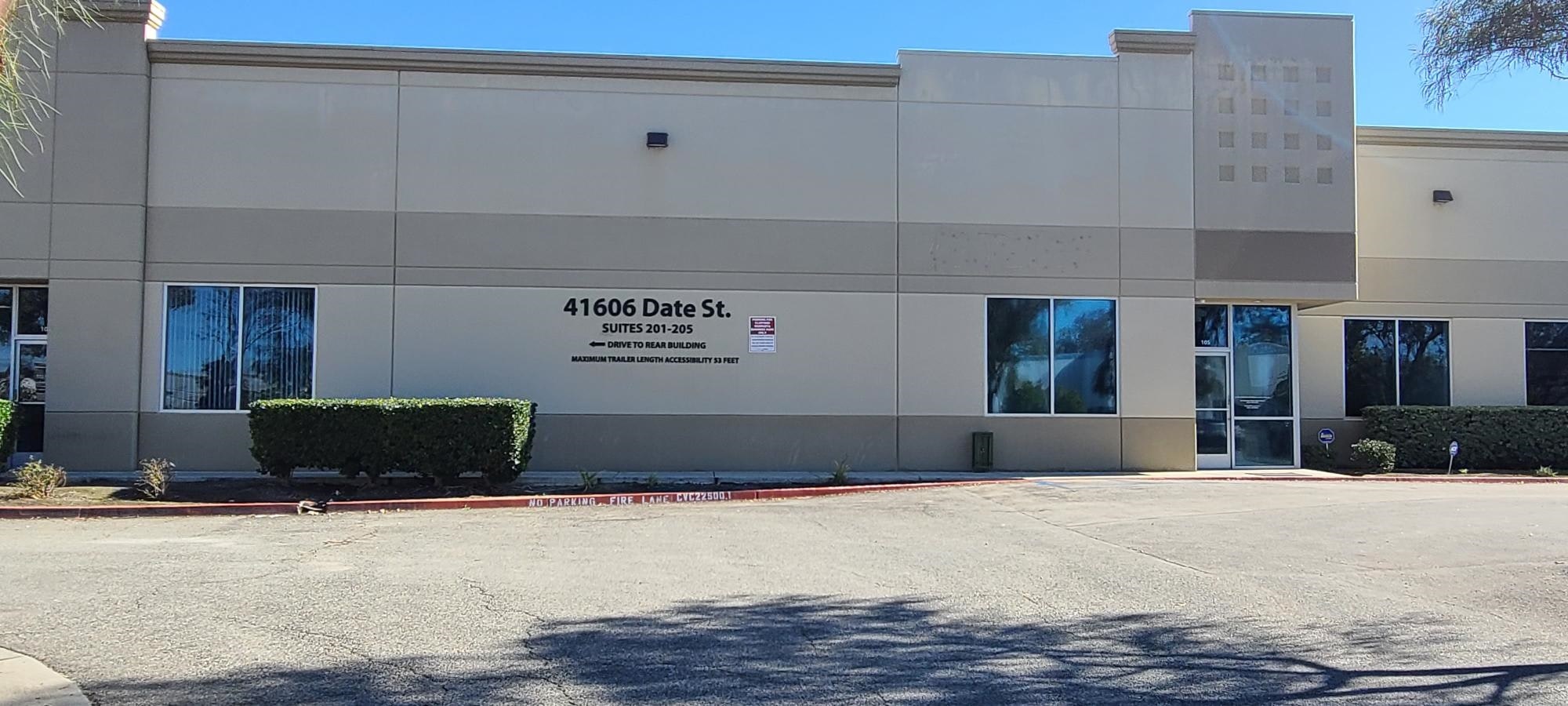 An exterior view of a building at 41606 Date St. with suites 201-205, including doors, windows, and a no-parking fire lane sign.