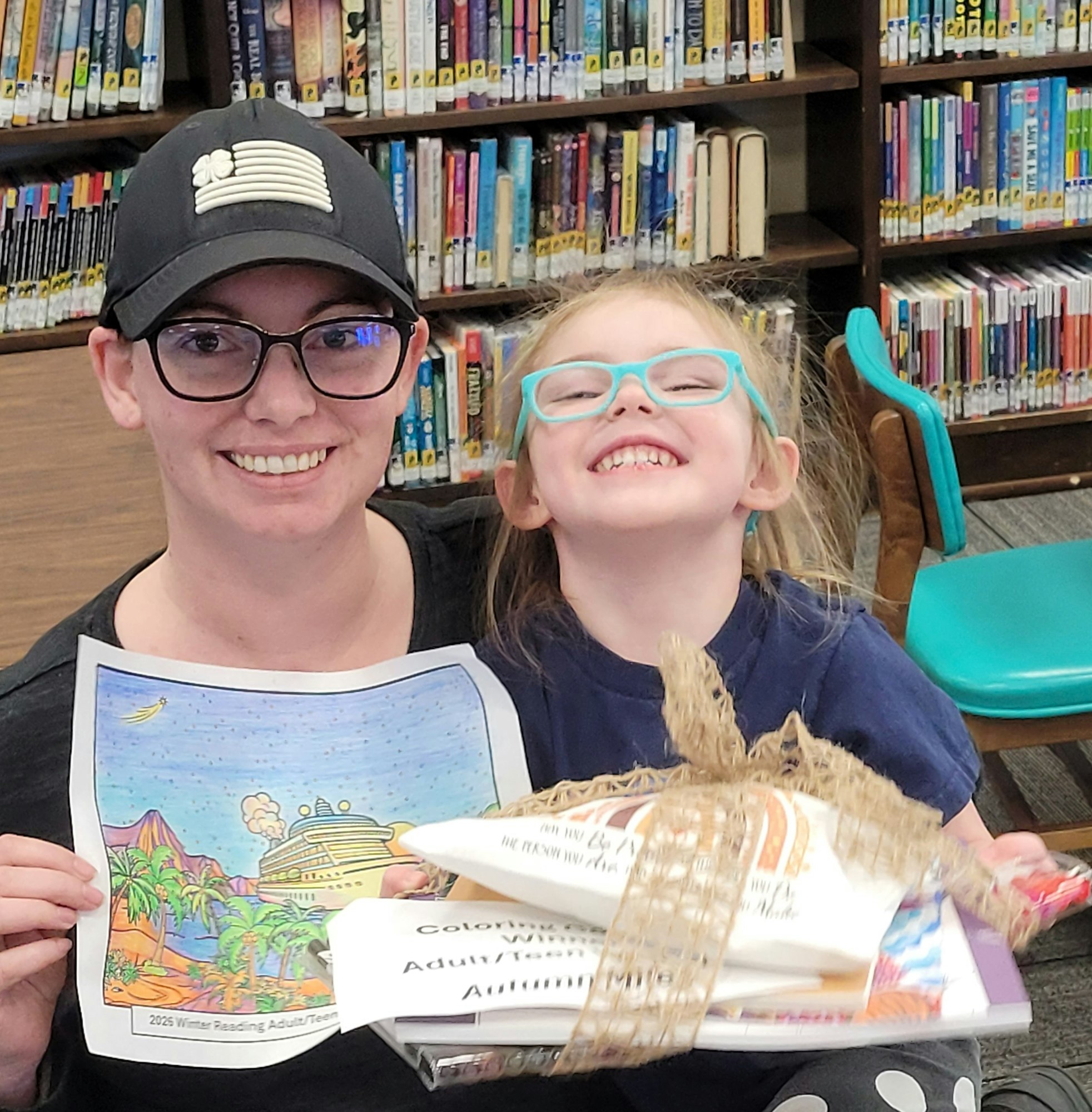 A smiling woman and child hold a colorful drawing and a prize bundle in a library, celebrating a reading program.