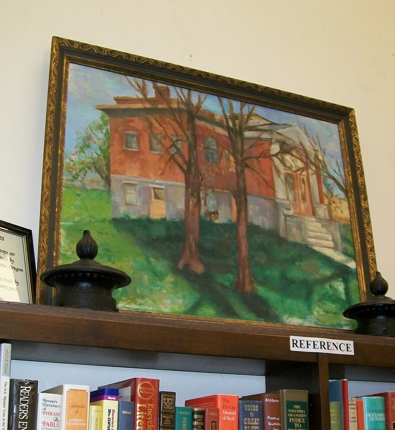 A painting of a red house with trees, displayed above a bookshelf filled with books, featuring a "REFERENCE" label.