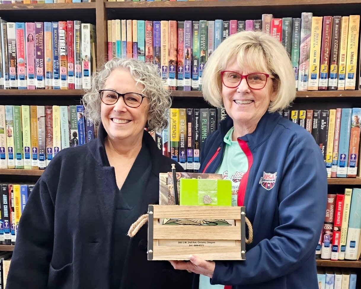 Two women smiling in front of a bookshelf, one holding a small wooden crate.