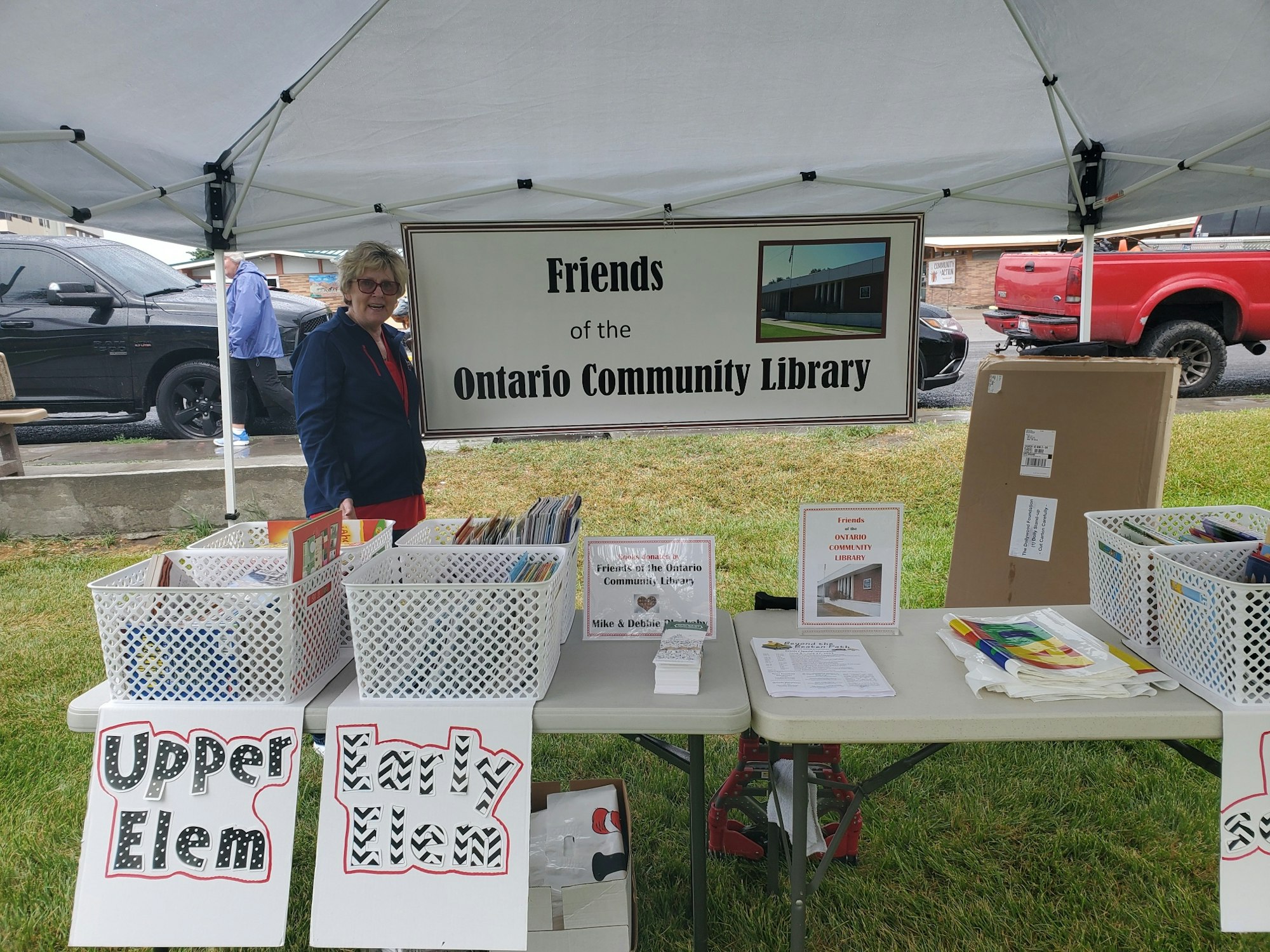 An outdoor stall with books labeled "Upper Elem," a sign saying "Friends of the Ontario Community Library," and a smiling person.