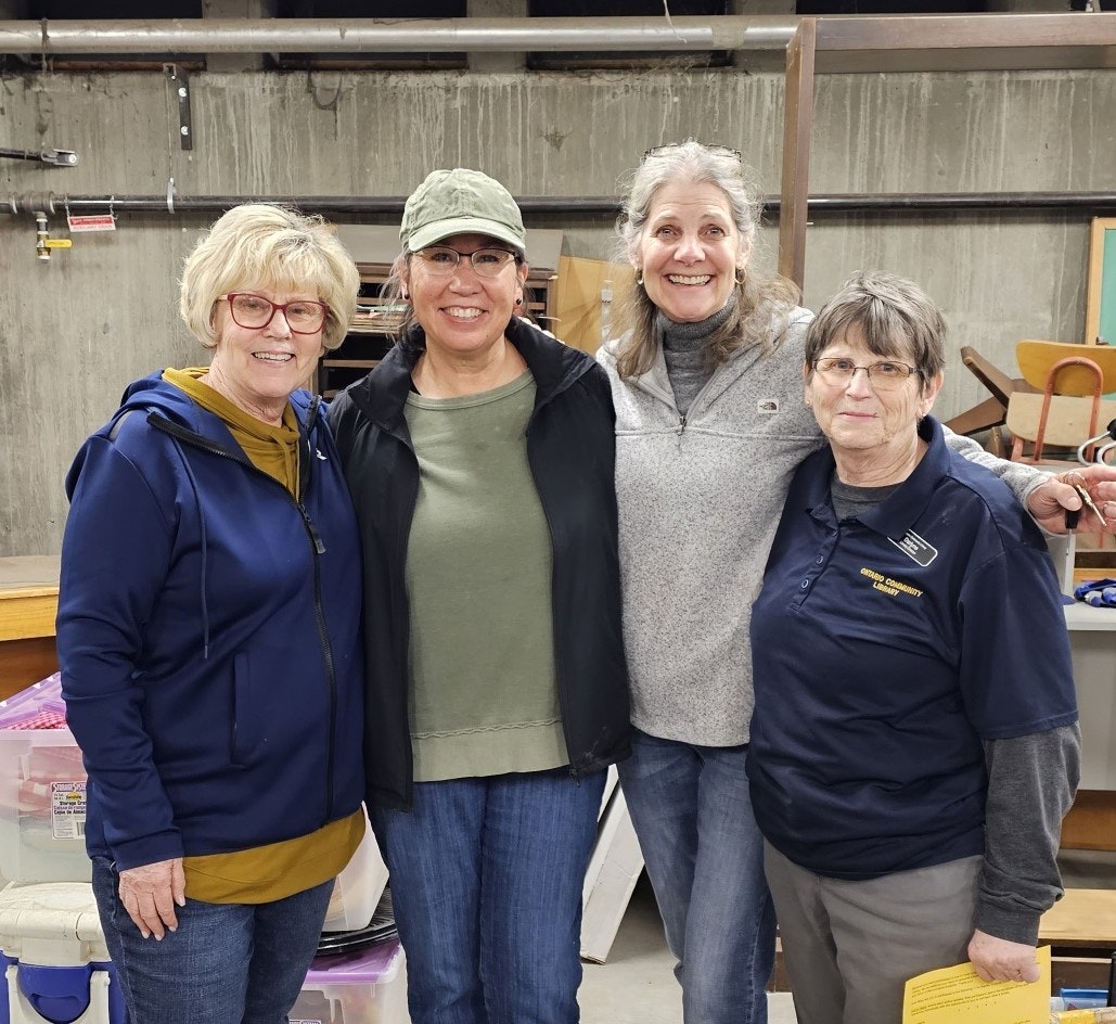 Four smiling women posing together in a workshop or garage setting.