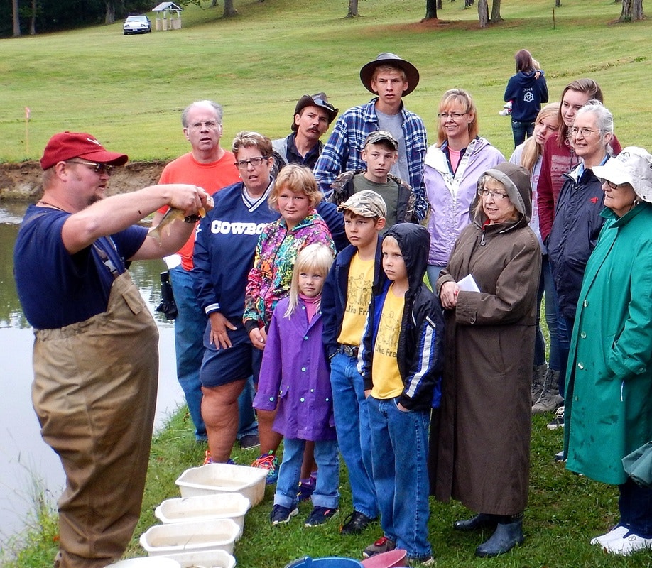 A group of people, including children and adults, gathered outdoors around a man demonstrating something.