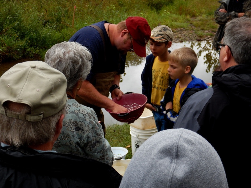 A group of people gathered outdoors, observing a man showing something in a bowl.