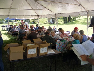 People gathered under a large tent, sitting at tables, eating and socializing outdoors with boxes and papers on tables.
