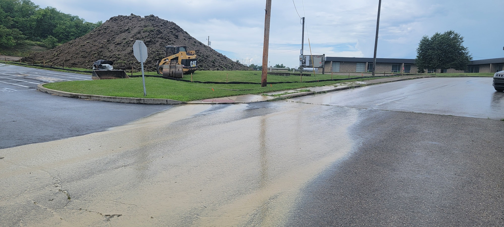 Wet pavement with muddy runoff, large dirt mound, construction vehicle, and overcast sky.
