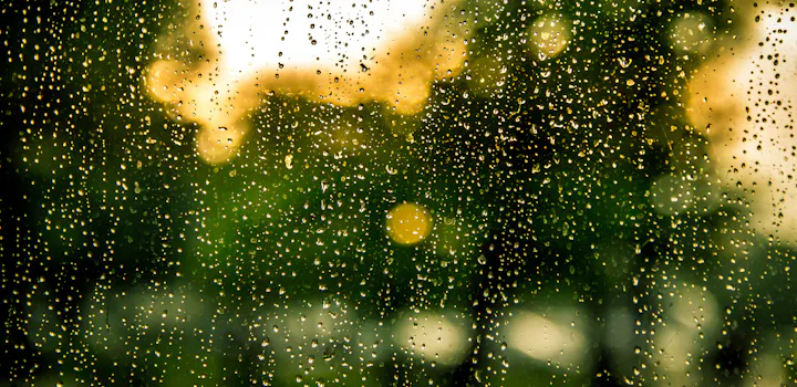 A close-up of raindrops on a window, with a blurred green and golden background, creating a soothing, dreamy atmosphere.