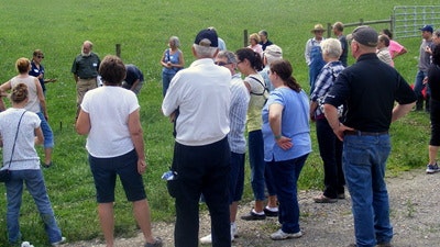 A group of people gathered outdoors, possibly for a tour or demonstration, facing someone speaking in a grassy area.