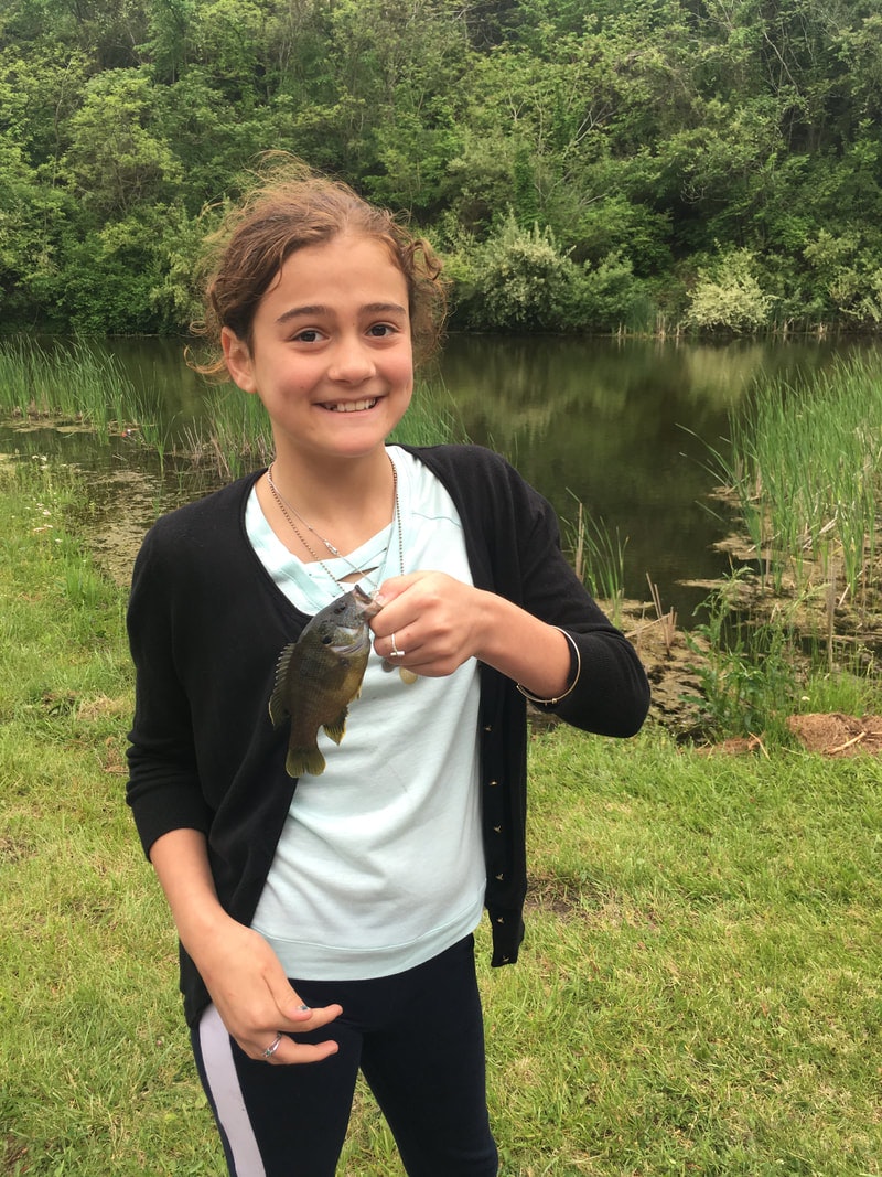 A person holding a small fish with a lake and greenery in the background.