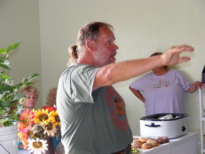 A man speaking indoors near a table with a slow cooker and pastries; sunflowers in the background.