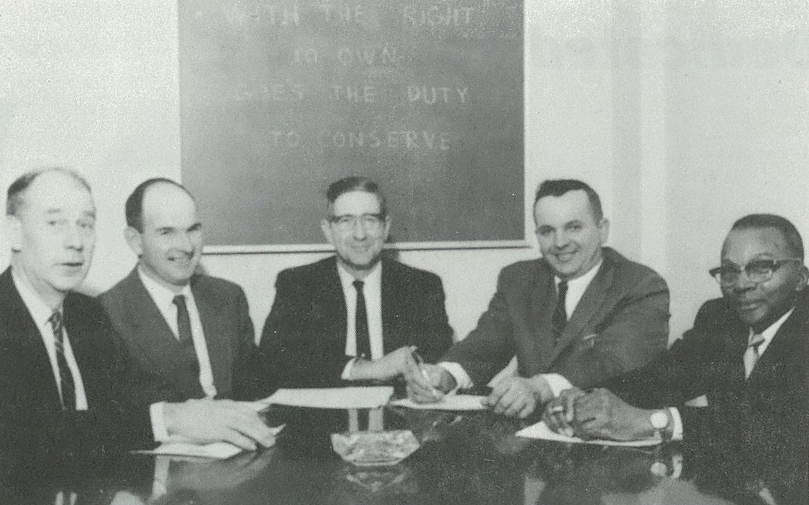 Five men in suits sitting at a table with papers, in front of a board with text.