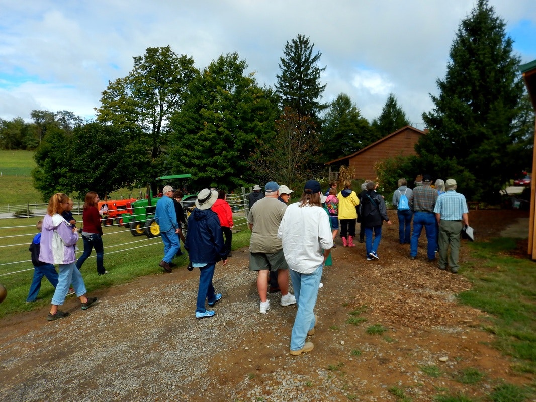 A group of people walking outside, near a farm with trees and a tractor in the background.