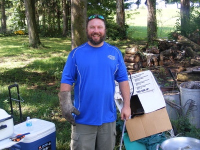 A man wearing a blue shirt and glove stands outdoors near boxes and a cooler, with trees in the background.