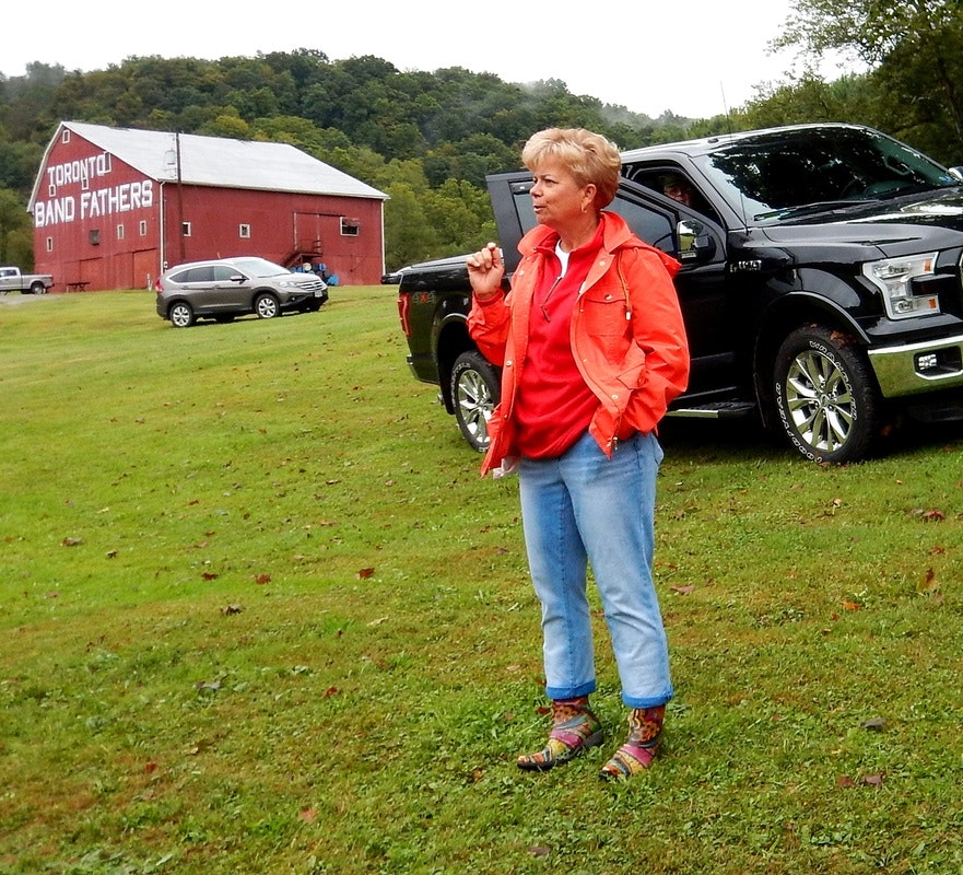 A person in an orange jacket stands on grass with a black truck; a barn labeled "Toronto Band Fathers" is in the background.