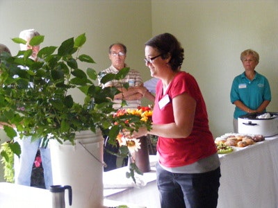 A woman arranges flowers at a table with snacks and a crockpot while others watch in a room.