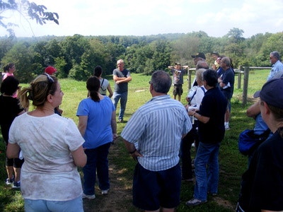 A group of people gathered outdoors, listening to a person speaking, with a scenic landscape in the background.