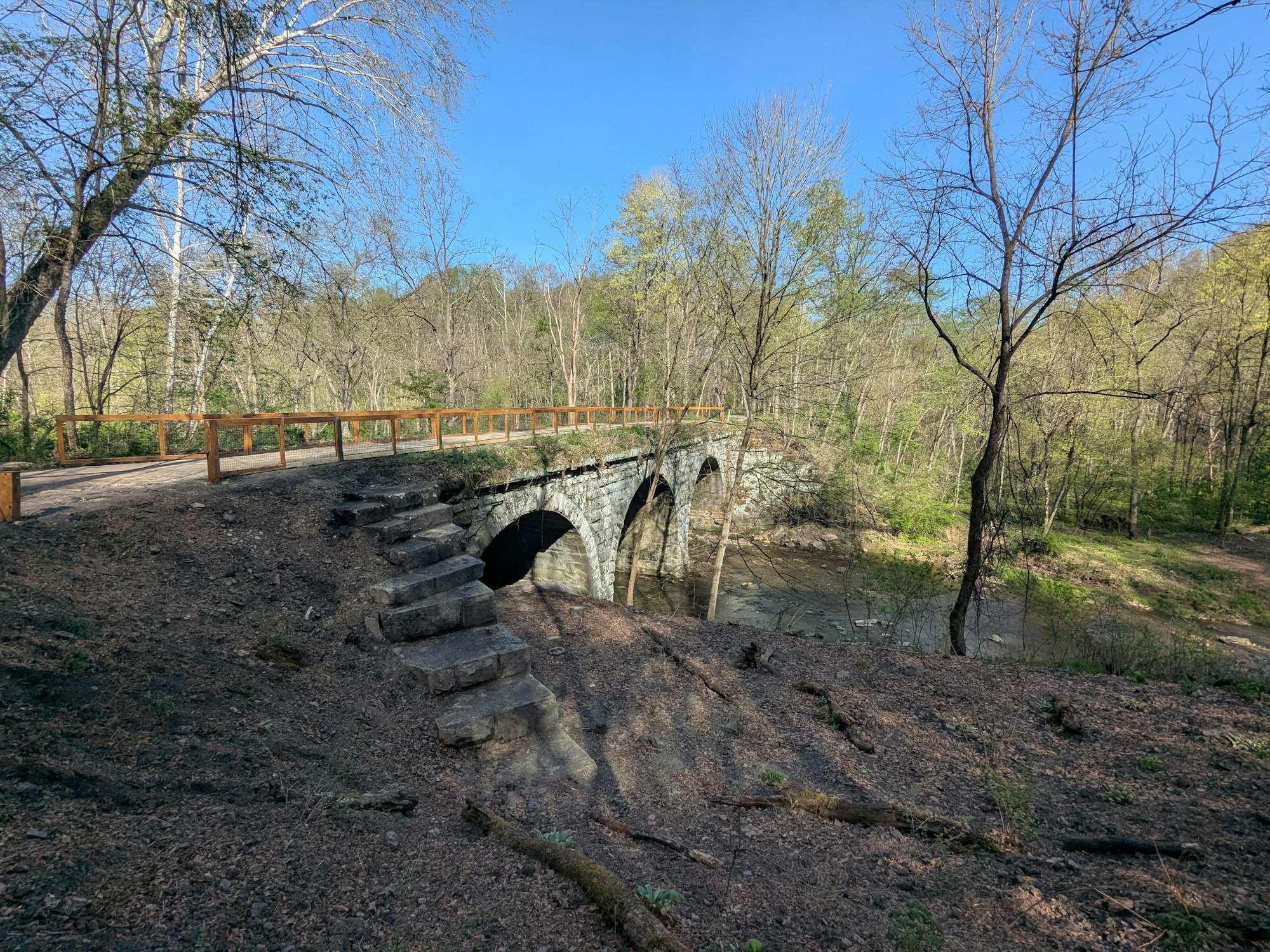 A stone bridge with arches over a creek in a forested area, under a clear blue sky.