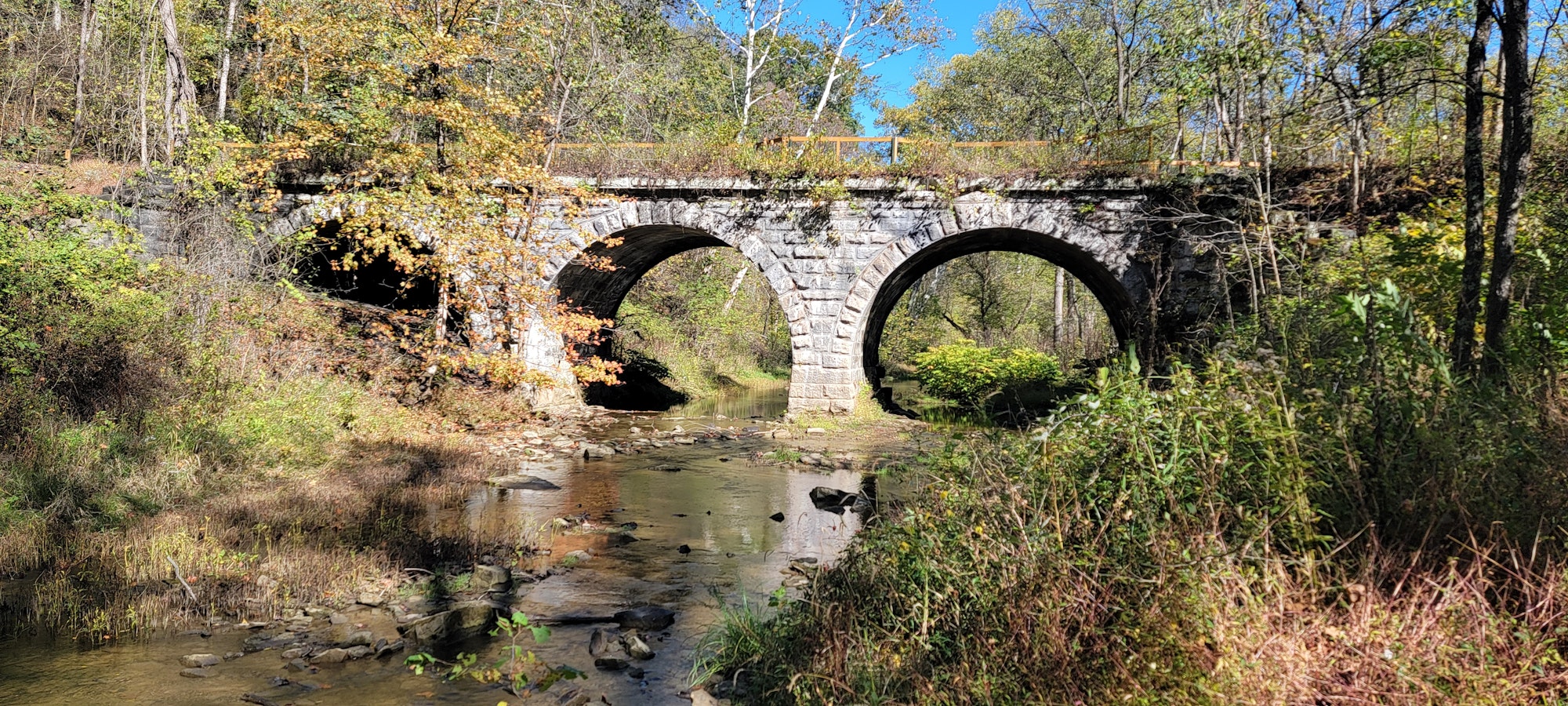 A stone arch bridge over a small stream surrounded by trees and foliage.