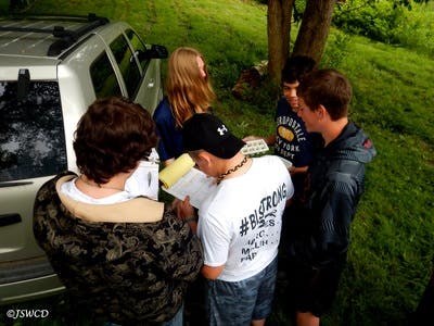 Five people standing near a car, looking down at papers and a carton of eggs outdoors.