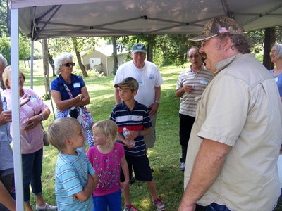 A group of people, including children and adults, are gathered under a canopy in a park or garden. They're casually dressed and talking.