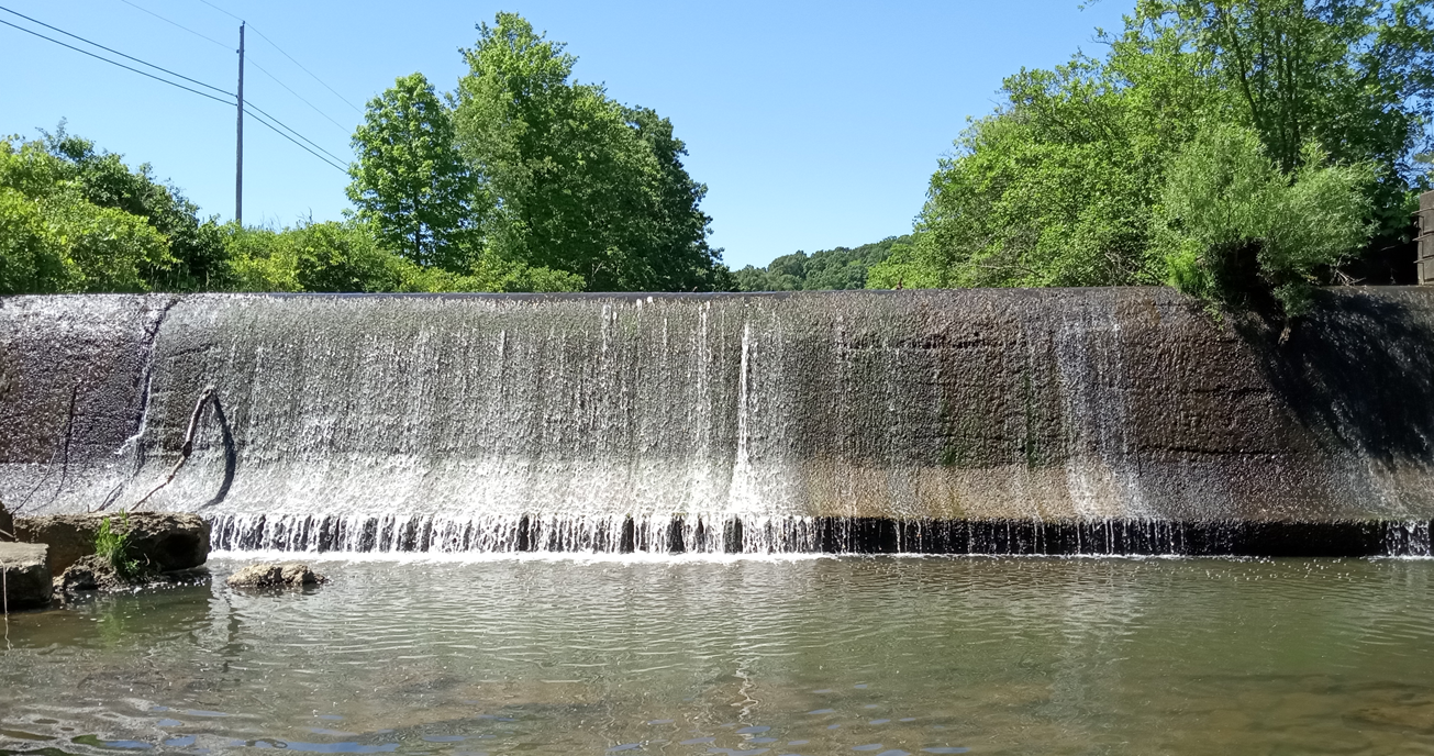 A small waterfall flows over a concrete wall into a calm river, surrounded by lush greenery and a clear blue sky.