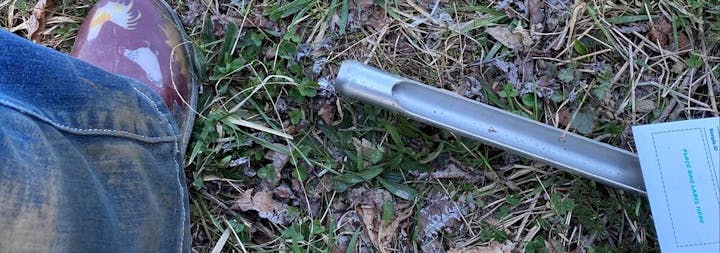 A person's leg in jeans and boots next to a soil sampling tool on grassy ground.