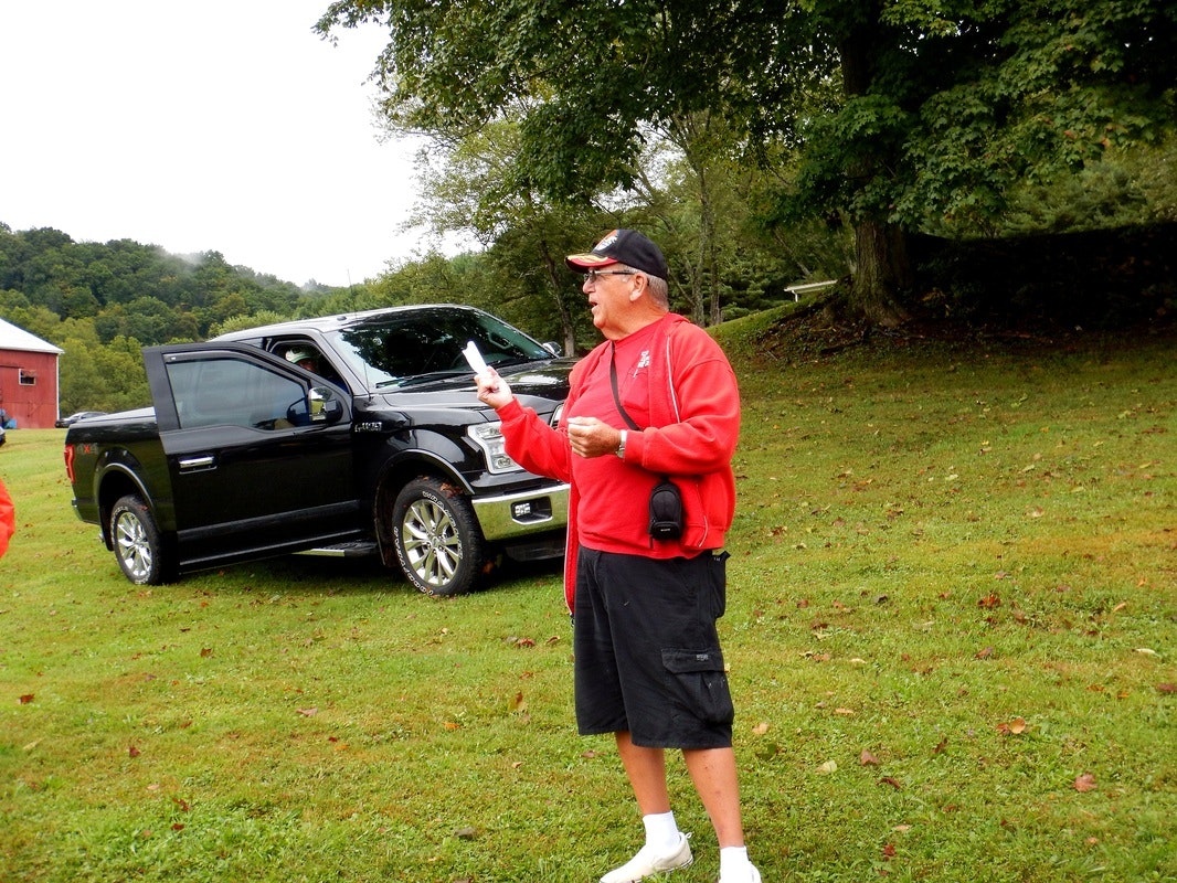 Man in a red jacket and cap holding papers, standing on grass beside a black pickup truck. Trees and a barn in the background.