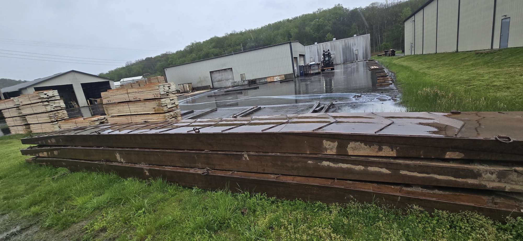 The image shows stacks of lumber and metal beams, with a wet surface, likely from rain, and storage buildings in the background.