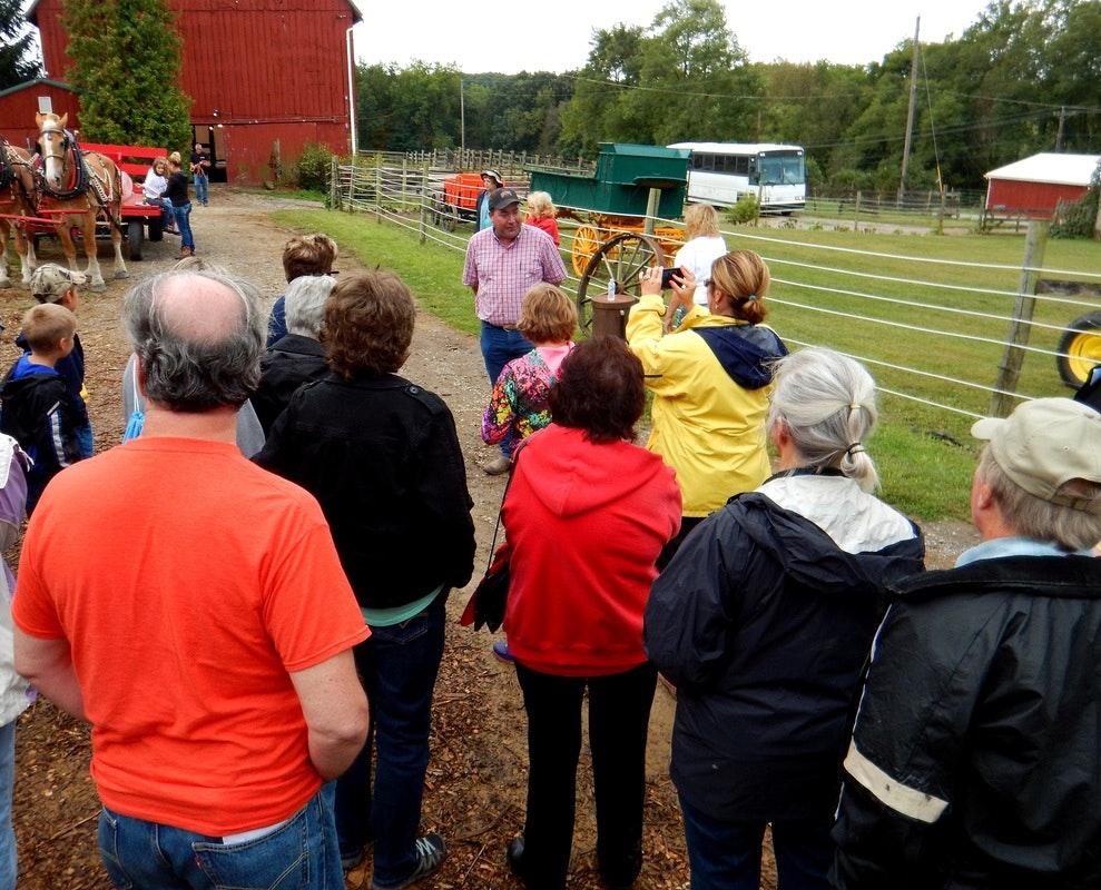 A group of people on a farm tour, listening to a guide near a red barn with horses and a wagon in the background.