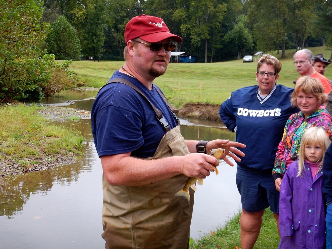 A man holding a fish stands near a stream, surrounded by a group of onlookers in an outdoor setting.
