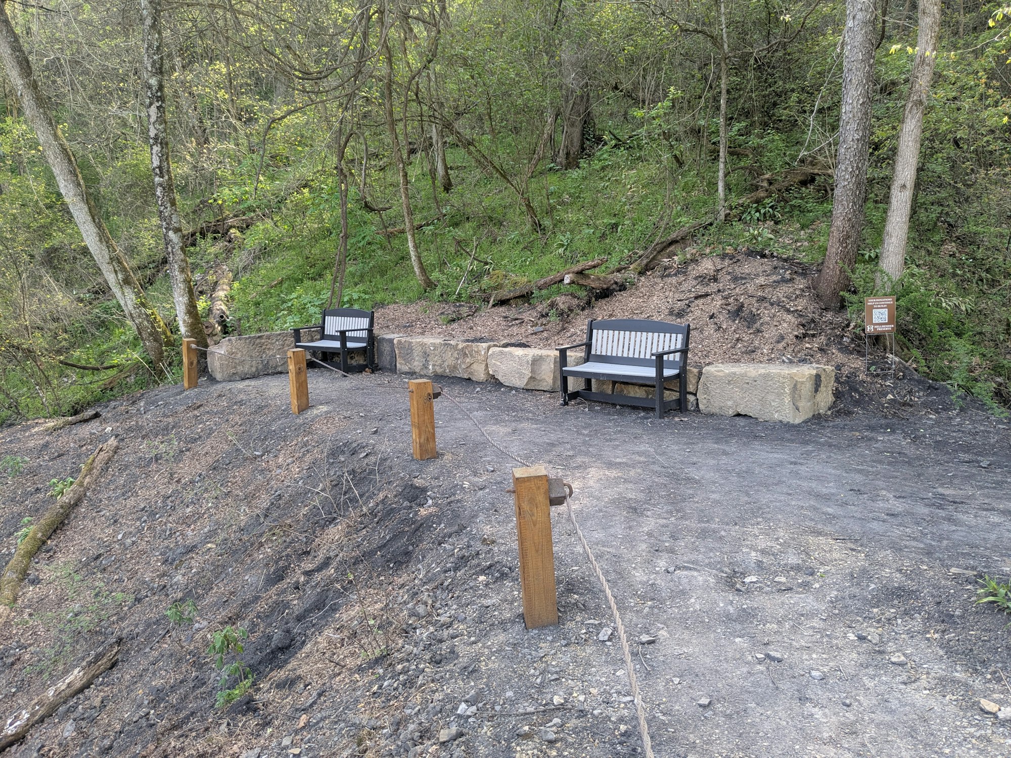 Two benches on a dirt path in a forested area, with wooden posts and rope nearby.